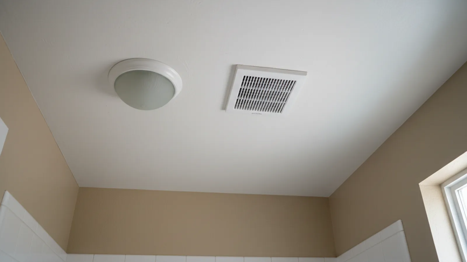 A plain white bathroom exhaust fan grille set flush in a painted drywall ceiling beside a ceiling light, the finished fan an inspector looks at after a bathroom fan installation