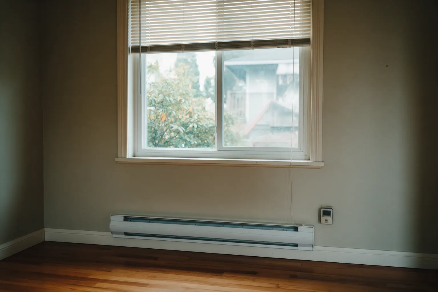 An electric baseboard heater under a window with a digital wall thermostat beside it